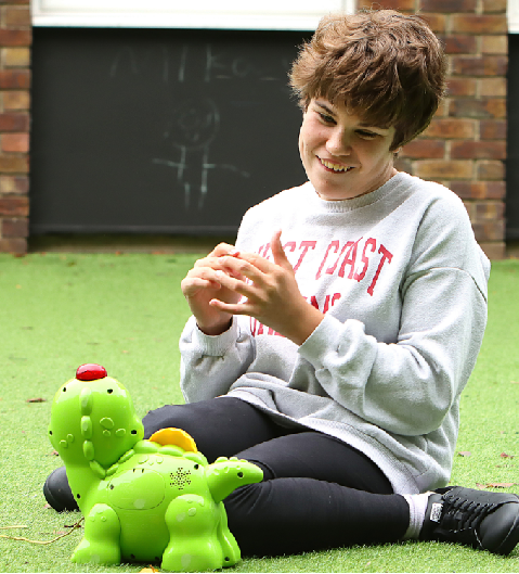 A photo of a young person sitting on the grass at our overnight respite unit for disabled children. The young person is looking at a dinosaur toy.