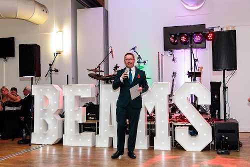 Beams parent, George Crockford has a microphone and is speaking to the guests attending the ball. there are white light up letters behind him that spell the word BEAMS