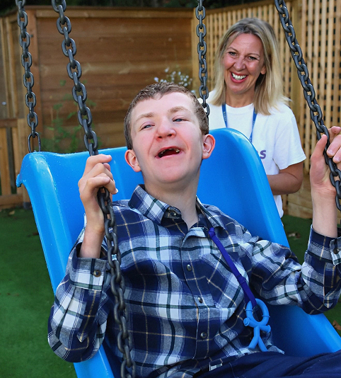 Photo shows a Support Worker called Tracey, pushing a blue swing with a young person who is smiling at our overnight respite unit, Dragon's Retreat.