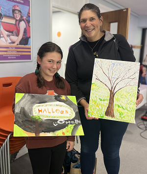 Photo shows a mum and daughter who has taken part in a artworkshop. They are holding a canvas each with a drawing of a tree.