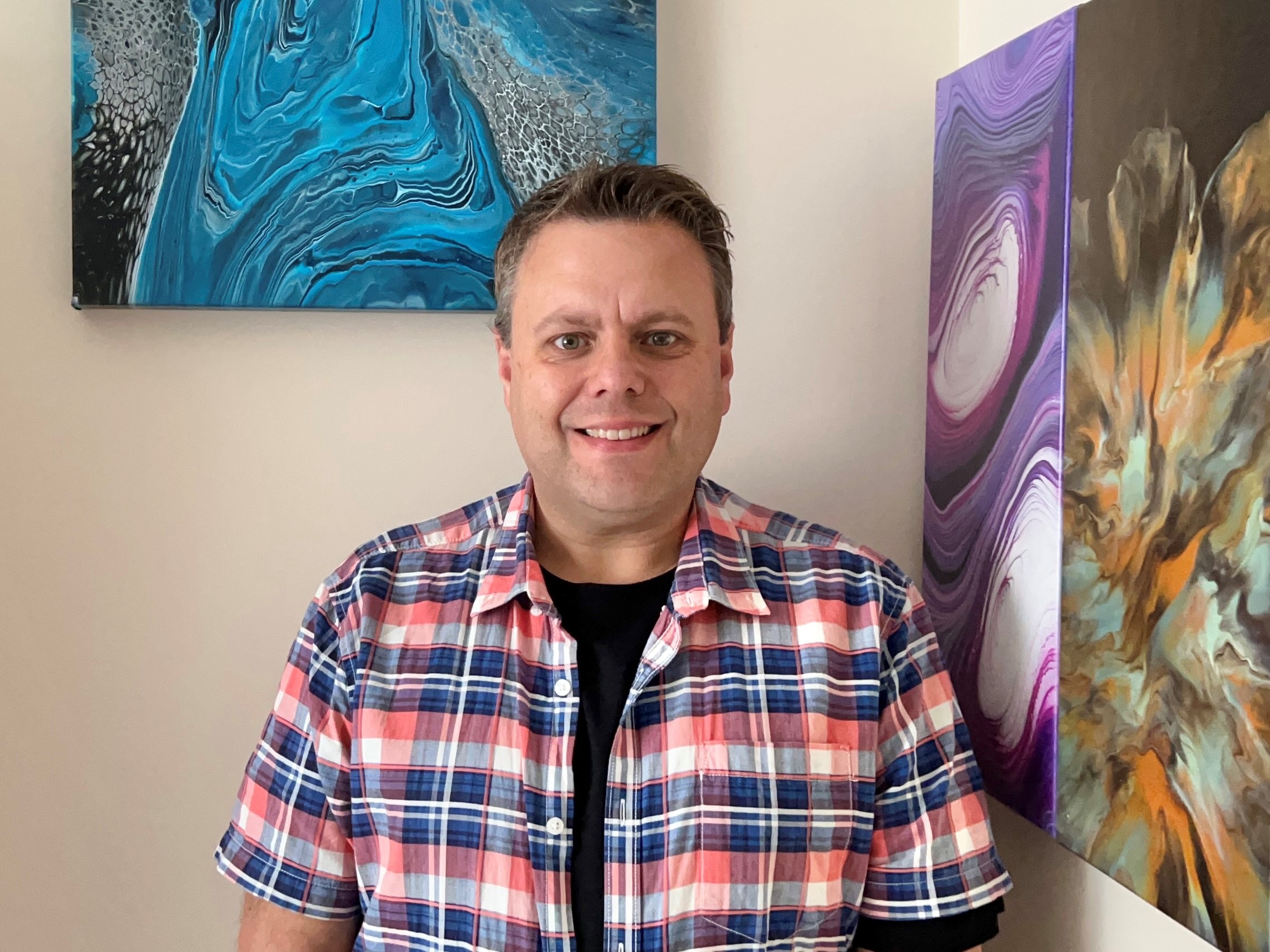 Image shows Mark Ratcliffe who is taking part in the our art auction, Inspired Expression. He is wearing a chequered shirt and standing by a blue piece of art, smiling.